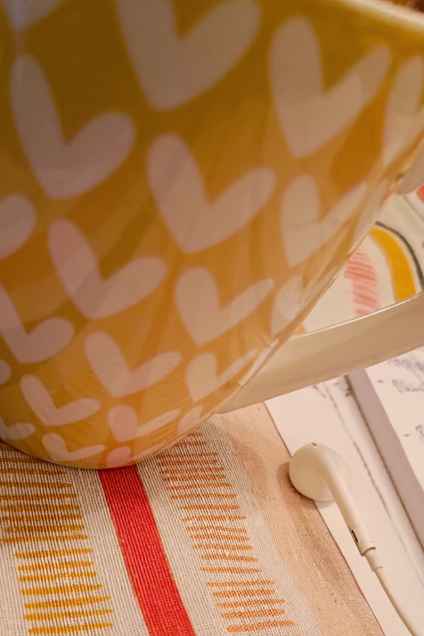 Kaffeetasse mit Herzchen vor gelbem Hintergrund auf bunter Tischdecke auf einem Arbeitstisch, 12 von 12
