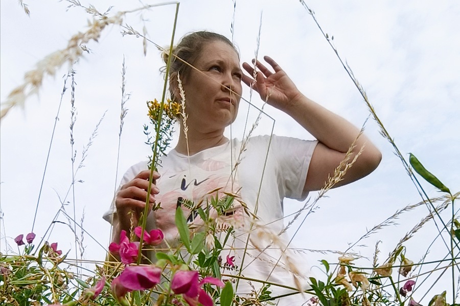 Inga mit Johanniskraut in der Hand inmitten blühender Sommerwildblumen, den Blick in die Ferne gerichtet – Sinnbild für die stille Stärke der Kräuterfrauen.